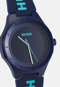 Reloj de silicona navy con una esfera redonda, que presenta una carátula oscura, agujas de horas y minutos blancas, y la marca "HUGO" en turquesa en la correa.