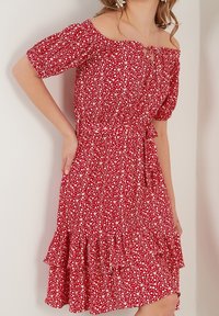 Femme portant une robe rouge à épaules dénudées avec un motif floral blanc, manches courtes, ourlet à volants et ceinture attachée à la taille, debout à l'intérieur.