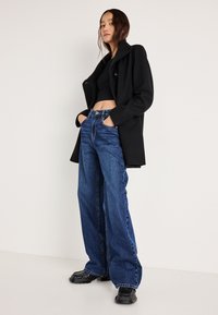 Čierny vlnený kabát s golierom, nosený cez skrátený čierny top. Modré široké džínsy s prednými vreckami a viditeľným šitím. Čierne topánky.