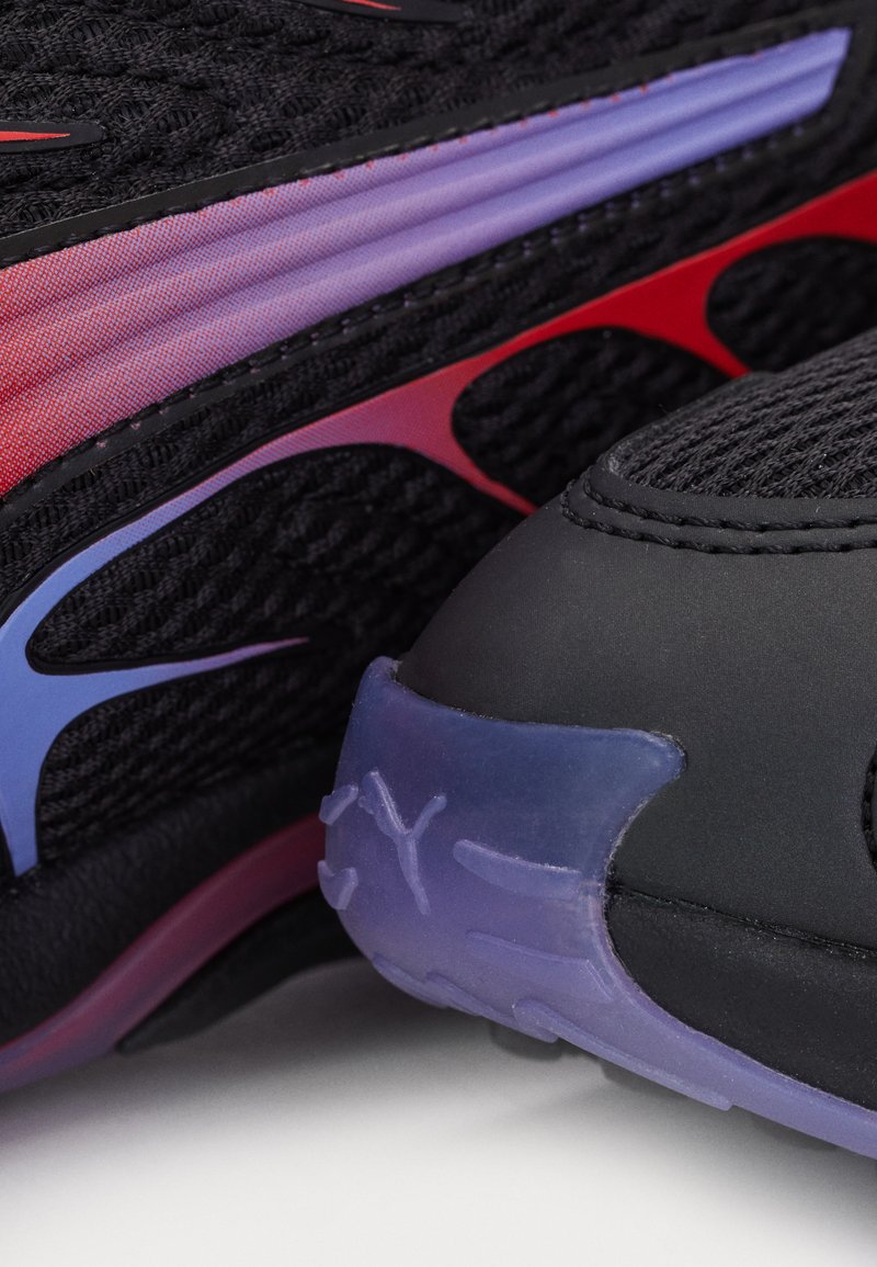 Zapatilla deportiva negra con parte superior de malla texturizada y acentos en rojo y púrpura, con una puntera negra brillante y un talón púrpura translúcido.