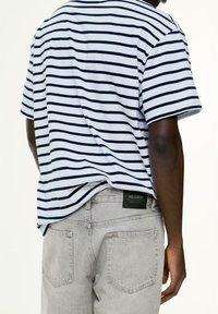 Person, der ein kurzärmliges, gestreiftes Hemd in Weiß und Marineblau trägt, das in hellgraue Jeans mit Gesäßtaschen und einem grünen Etikett gesteckt ist.