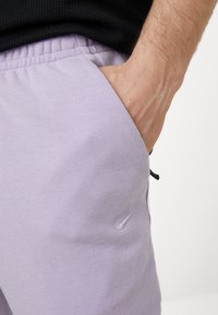 Light purple sweatpants with elastic waistband, side pocket, and a small embossed Nike logo on the left thigh. Smooth fabric texture.