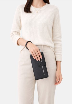 Donna con pantaloni beige e maglione bianco che tiene una pochette da polso nera con cerniera e cinturino con bottone a pressione all'altezza della vita.