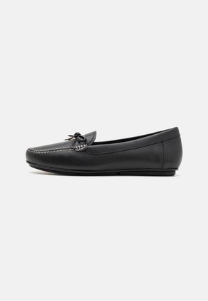 Mocassin en cuir noir avec des coutures blanches, détail de nœud et semelle en caoutchouc plate, présenté de côté sur fond blanc.