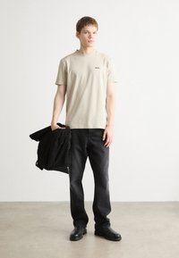 Beige bomulds t-shirt med et lille sort logo, parret med sorte denim jeans og en sort jakke i hånden. Glat, afslappet design.