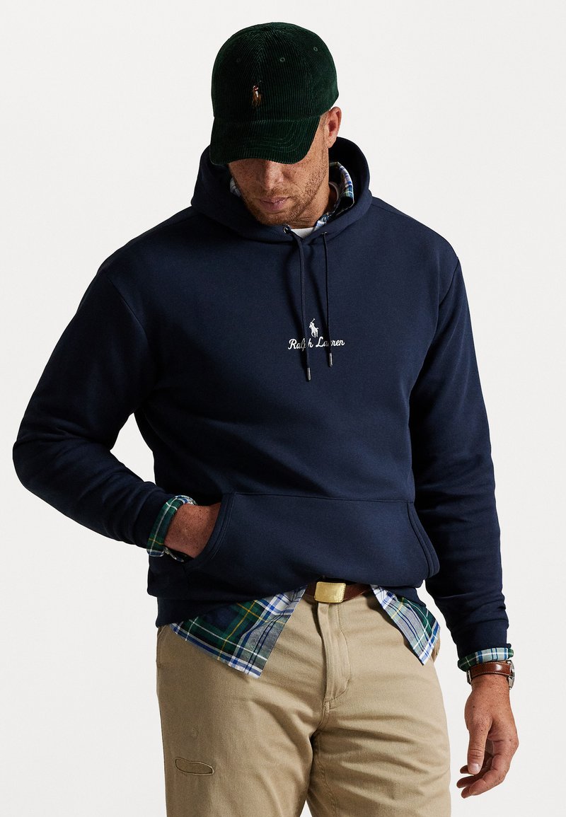 Uomo che indossa un hoodie blu navy con tasca frontale sopra una camicia a quadri verde, pantaloni beige, berretto verde e orologio da polso, in piedi su uno sfondo bianco.