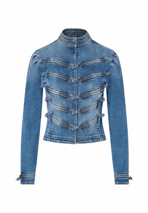 Femme portant une veste en jean ajustée avec des détails de boucles, un jean taille haute, des bottines marron et des bracelets dorés, debout les mains dans les poches.