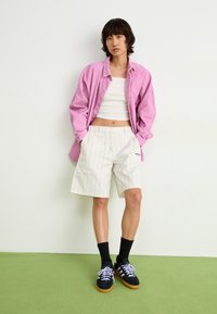 Model wearing a pink corduroy shirt, white striped shorts, and black sneakers with light purple laces, standing on a green floor.