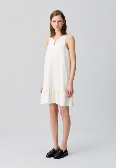 Vestido blanco sin mangas con escote en forma de ojal y dobladillo con volantes, combinado con mocasines de cuero negros con un acento dorado.