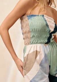 Combinaison sans bretelles avec un corsage froncé, en tissu texturé aux motifs abstraits verts, beiges et bleus. Poches latérales visibles.