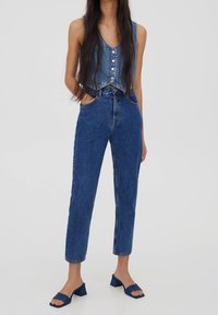 Denim crop top s knoflíkovým zapínáním, kombinovaný s modrými džíny s vysokým pasem. Modelka má na sobě modré vysoké sandály. Čistý, neformální design.