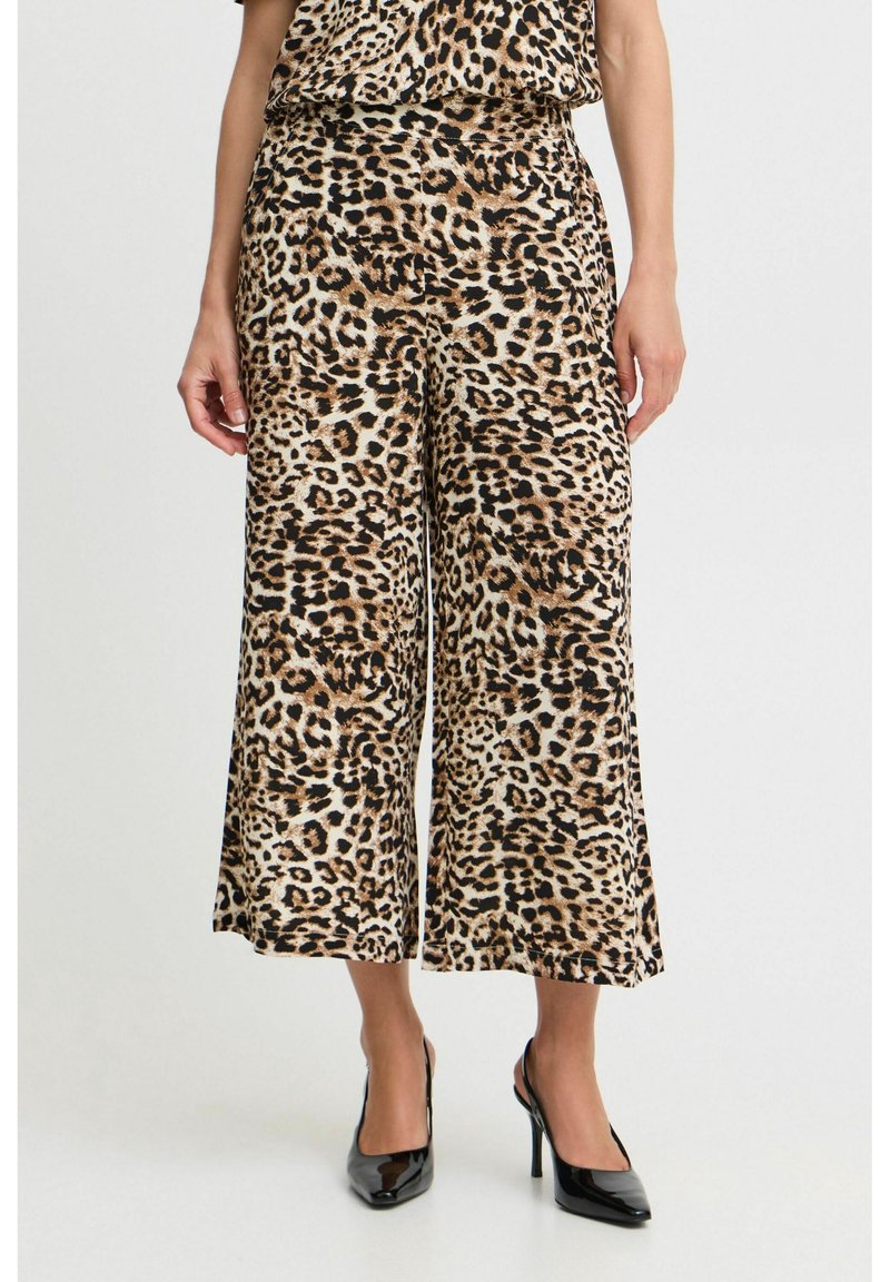 Femme portant un pantalon large coupé à l’imprimé léopard et un haut assorti, associé à des chaussures à talons hauts pointues noires, debout sur un sol blanc.