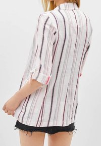 Vit skjorta med vertikala ränder i rosa och marinblått, uppvikta ärmar och krage. Tyget verkar lätt och har en lätt texturerad yta.