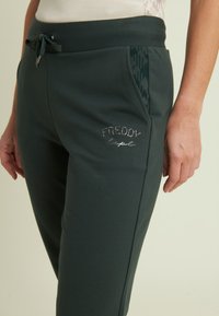 Jogging in verde scuro realizzati in materiale morbido, con vita regolabile mediante cordino, tasche laterali con dettagli a fantasia e logo glitterato.