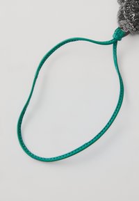 Zöld, fonott bőrszíj sima felülettel, egy hurokkal és csomózott részlettel, amely egy dekoratív szürke virágmotívum közelében található.