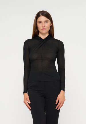 Femme aux cheveux bruns et raides porte un haut noir ajusté à manches longues avec un col croisé et un pantalon noir, debout sur fond blanc.