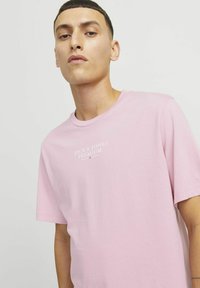Šviesiai rožinė medvilninė marškinė su apvalia apykakle ir trumpomis rankovėmis. Priekyje yra balta spausdinta prekinio ženklo užrašas "JACK & JONES PREMIUM."