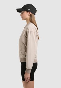 Jeune femme en veste bomber beige, short de cyclisme noir et casquette de baseball noire, debout de profil devant un fond uni.
