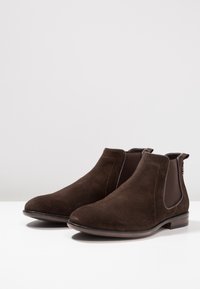 Bottines en daim marron avec panneaux élastiques latéraux, bouts ronds et semelles en caoutchouc marron foncé. Des coutures discrètes ajoutent du détail.