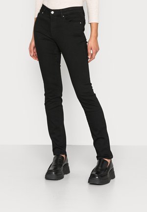 Jeans negros ajustados con una cintura de altura media, bolsillos delanteros y una textura de mezclilla suave. Combinados con zapatos negros tipo slip-on de suela gruesa.