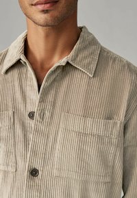 Chemise en velours côtelé beige avec col, deux poches avant et fermeture à boutons. La texture est côtelée avec une finition douce et veloutée.