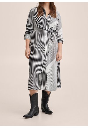 Femme portant une robe à boutons à rayures verticales noir et blanc nouée à la taille, avec des bottes de cow-boy noires arrivant au mollet sur un fond beige.