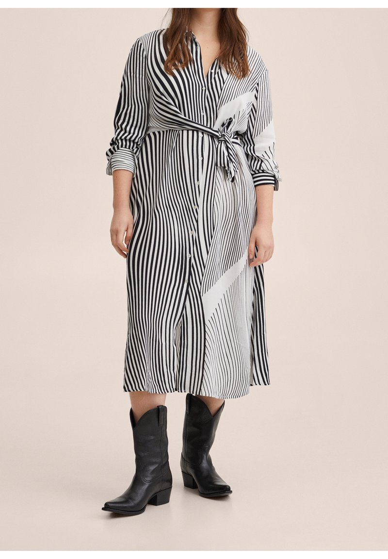 Femme portant une robe à boutons à rayures verticales noir et blanc nouée à la taille, avec des bottes de cow-boy noires arrivant au mollet sur un fond beige.