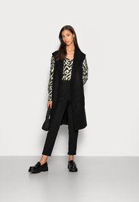 Colete longo preto por cima de uma blusa estampada em preto e branco, calças pretas slim e sapatos de plataforma pretos. Inclui uma mala preta texturizada.