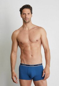 Modré pánské boxerky s hladkým povrchem, s kontrastním černým páskem se stříbrným logem "yamamay". Model stojí na neutrálním pozadí.