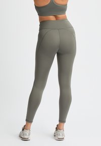 Leggings a vita alta di colore verde oliva realizzati in tessuto elasticizzato, con dettagli delle cuciture lungo le gambe, abbinati a scarpe da ginnastica argentate.
