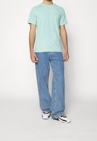 Světlý modrozelený tričko s krátkým rukávem a decentním logem na hrudi, spárované s volnými světle modrými džíny.