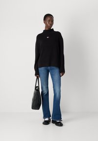 Svart ribbet genser med høy hals, blå flagrende jeans, svarte platåsko og en teksturert svart totebag med geometrisk design.