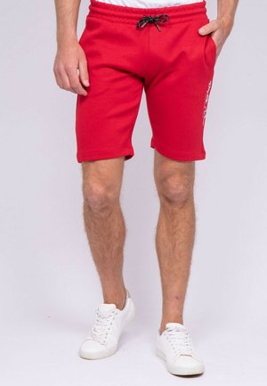 Homme portant un short rouge vif à cordon de serrage et des baskets blanches, debout avec une main dans la poche sur un fond uni.