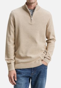 Beige gebreide pullover met een kwart-rits, ribgebreide kraag en gestructureerde stof. Gecombineerd met blauwe spijkerbroek.