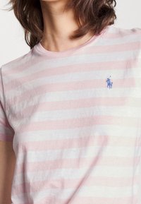 Kortärmad randig t-shirt i ljusrosa och vitt, gjord av mjukt tyg, med en liten blå broderad logotyp på vänster bröst.