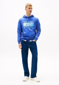Blaues Baumwoll-Hoodie mit grafischem Design, verstellbare Kapuze und Kängurutasche, kombiniert mit blauen Jeans und weißen Sneakern.