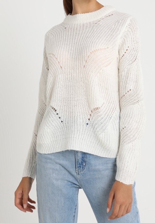 Femme portant un pull blanc à maille lâche avec des motifs ondulés, associé à un jean en denim bleu clair, sur un fond uni.