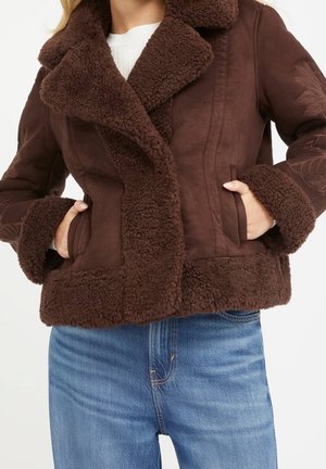 Woman wearing a brown shearling jacket with thick collar and cuff trim, paired with blue jeans, hands in jacket pockets.