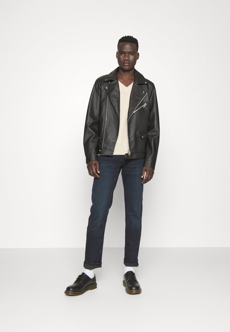 Junger Mann in schwarzer Lederjacke, beigem V-Ausschnitt-Pullover, dunklen hochgekrempelten Jeans, weißen Socken und schwarzen Schuhen, der vor einem schlichten weißen Hintergrund steht.