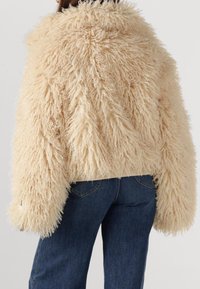 Fuzzy beige faux fur jacket with a textured, shaggy exterior and wide sleeves, paired with dark blue jeans. Visible stitching detail.