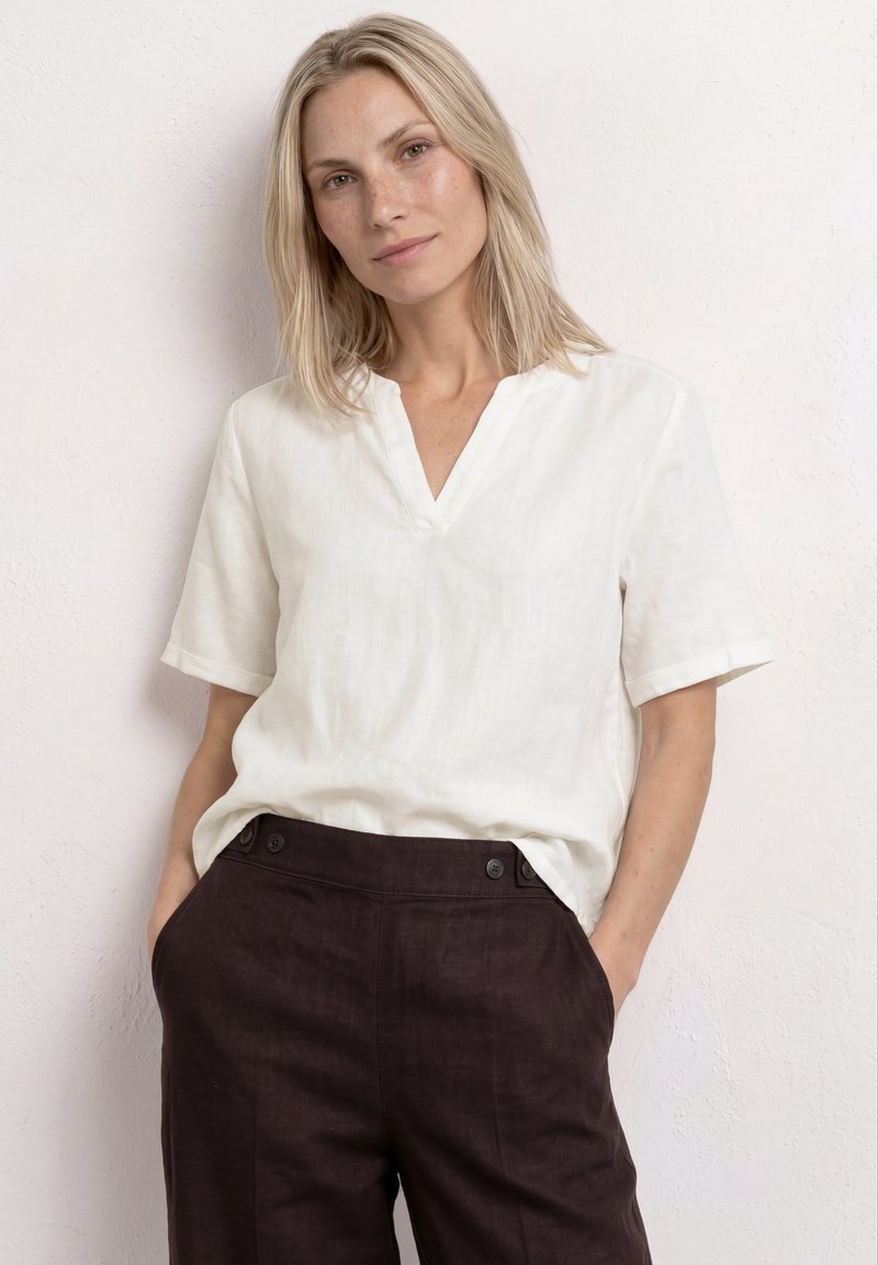 Femme blonde portant un haut blanc à manches courtes en V et un pantalon taille haute foncé, les mains dans les poches, debout contre un mur clair.