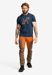 Marinblå t-shirt med en orange logotyp, kombinerad med orange och bruna flerficksbyxor, samt svarta snörda stövlar.