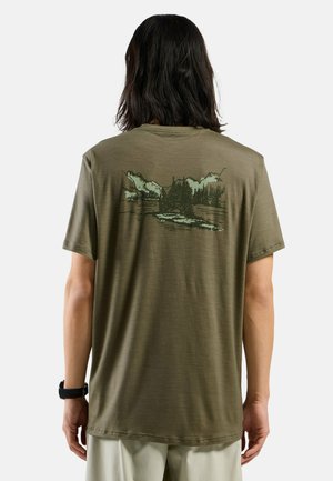 Person with shoulder-length dark hair wearing an olive green t-shirt with a mountain and forest graphic on the back.