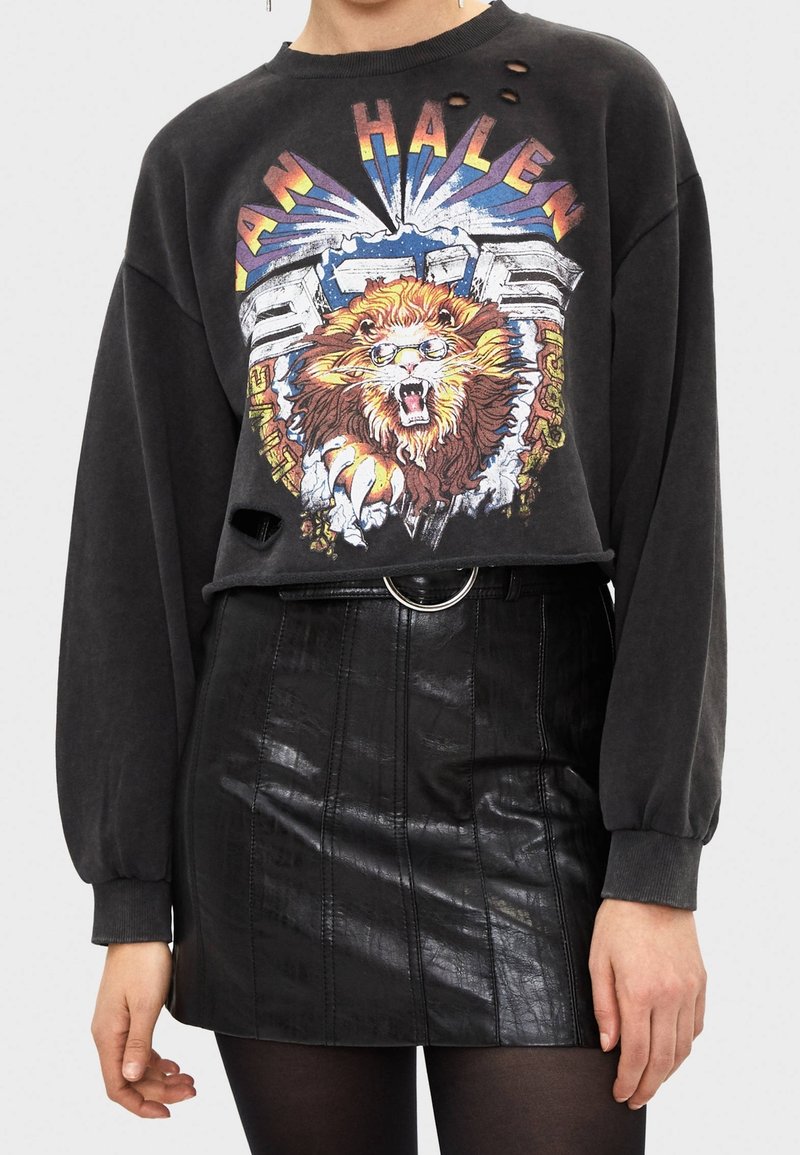 Svart, kort sweatshirt med grafik av ett lejon, med slitna hål. Kombinerad med en glänsande svart minikjol med vertikala paneler.