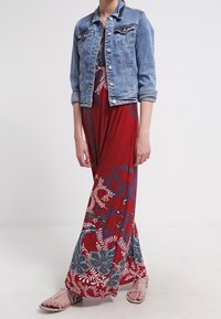 Veste en jean, robe longue rouge à motifs floraux bleus et blancs, et sandales argentées. La tenue présente un design fluide et une coupe décontractée.