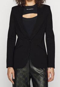 Černý přizpůsobený blazer s vykrojeným designem v oblasti hrudníku, s elegantními klopami, bočními kapsami a zapínáním na knoflík.