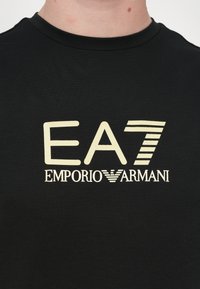 T-shirt preta de gola redonda com uma grande impressão gráfica bege de "EA7 EMPORIO ARMANI," com diferentes larguras de linha e um acabamento de tecido texturizado.