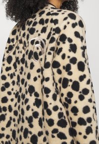 Beige fleecová bunda s černým leopardím potiskem, kulatým výstřihem a ozdobnou stříbrnou výšivkou na zadní straně. Měkká textura.