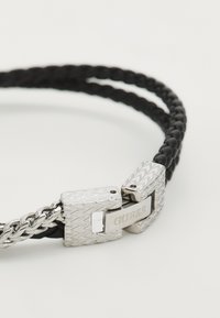 Svart flettehåndleddsarmbånd i lær med en sølvmetallklemme som har et sikksakkmønster og gravert logo. To sammenflettede bånd skaper tekstur.