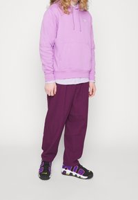 Ljuslila hoodie med en framficka, lagd över en randig skjorta. Löst sittande mörklila byxor och svarta sneakers med färgglada accenter.